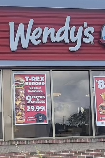 The exterior of a Wendy’s restaurant with a window advertisement showing “T-REX BURGER
Go Big or Go Extinct.
$29.99” with a picture of a giant 9-patty hamburger accompanying the caption.
